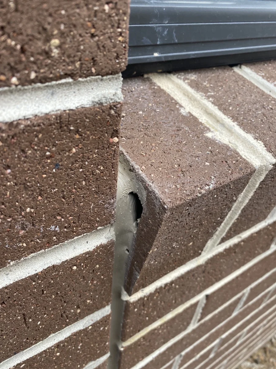 Close-up of a brick wall corner with a missing brick, creating a hole. The bricks are reddish-brown, with a black window sill above.