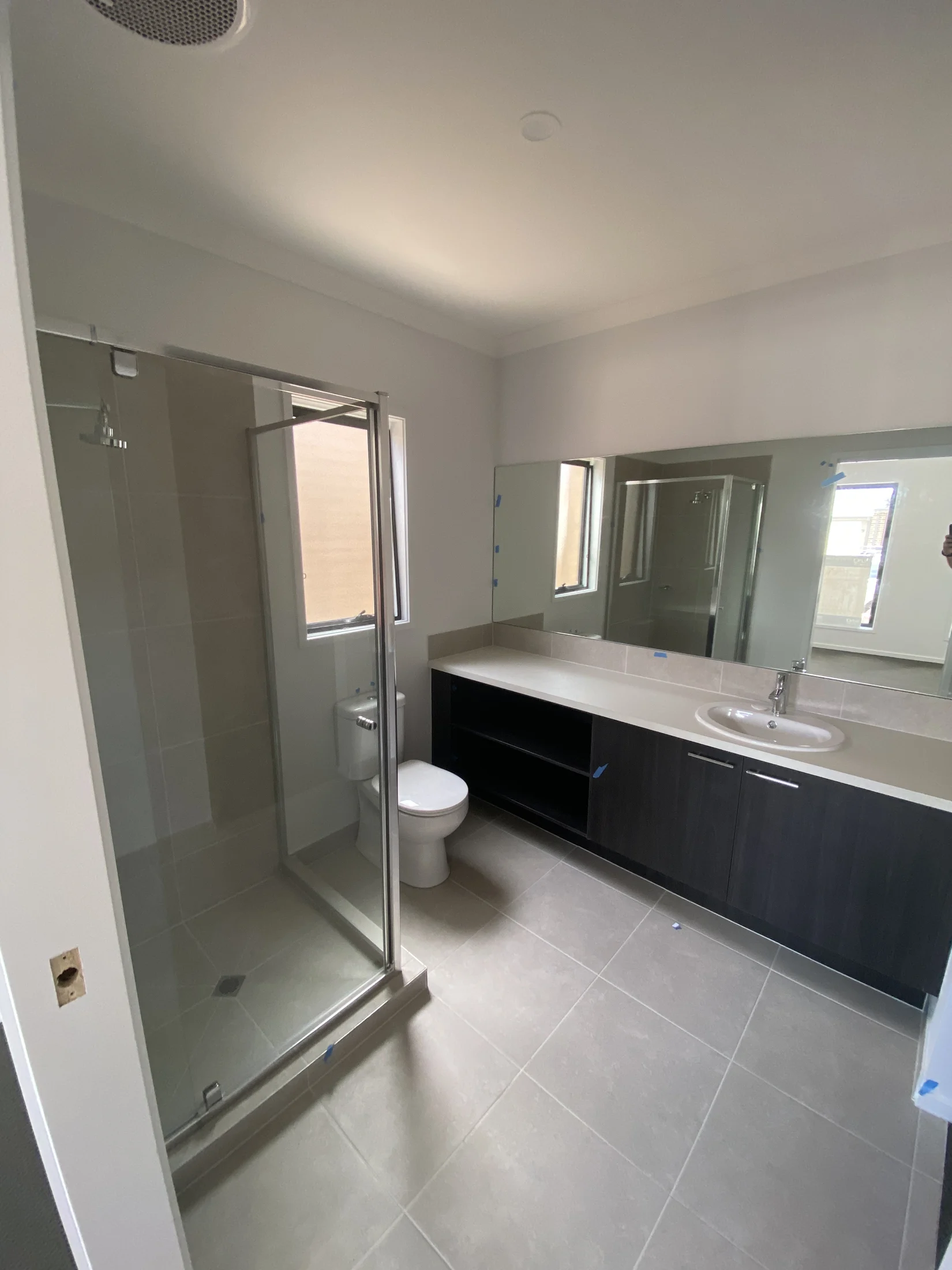 Modern bathroom with a glass shower, white toilet, and a dark wood vanity with a sink. Large mirror above, gray tiles, bright lighting.