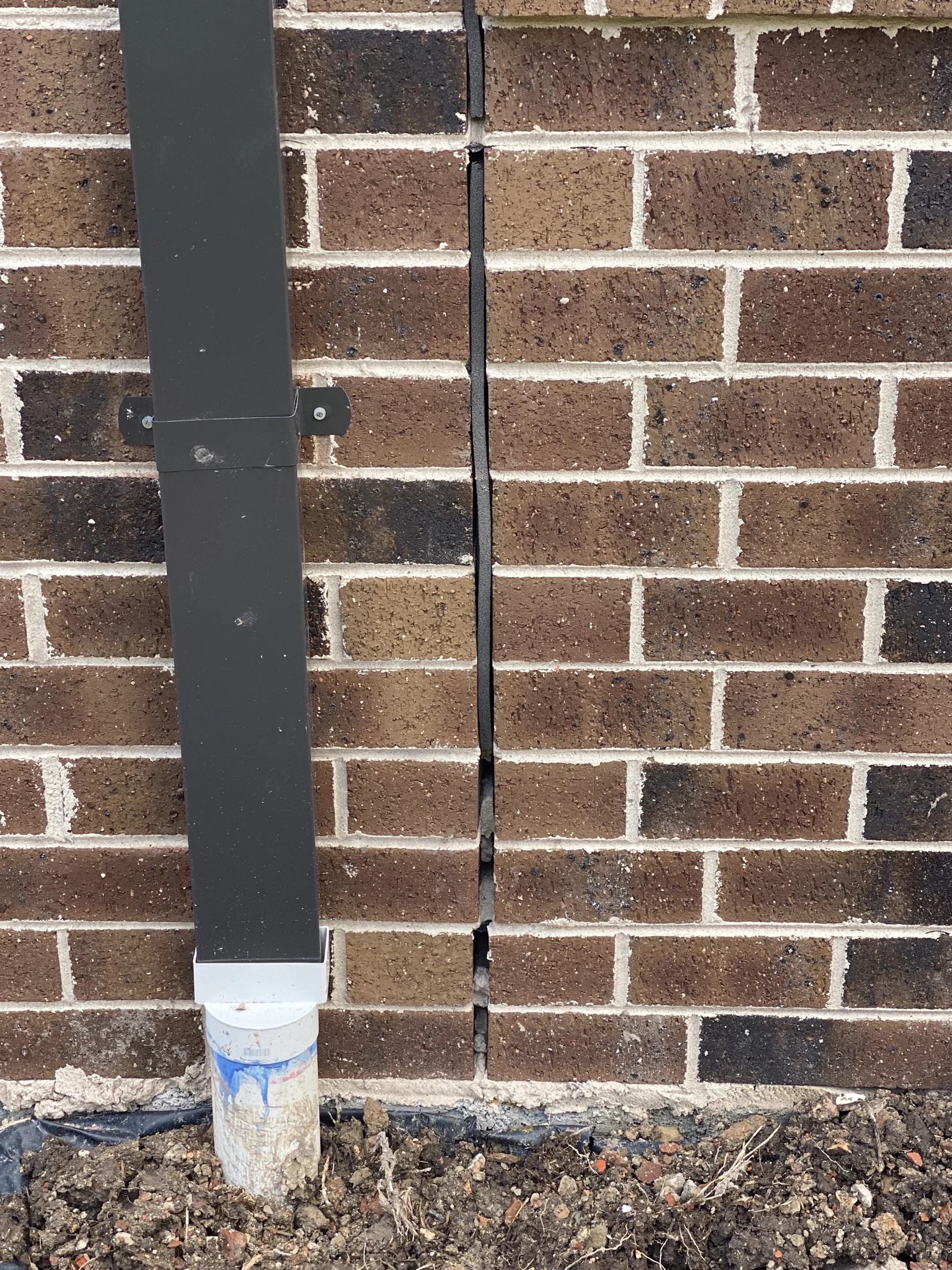 Brick wall with a vertical crack and attached downspout leading to PVC pipe. Soil and debris are at the base. Brown bricks with white mortar.