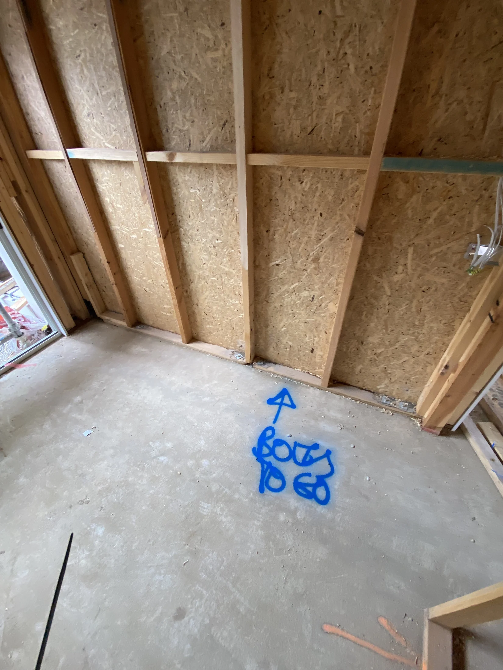 Wooden framed wall in an unfinished room with a cement floor. Blue spray paint reads "Bolts to go" with an arrow, indicating direction.