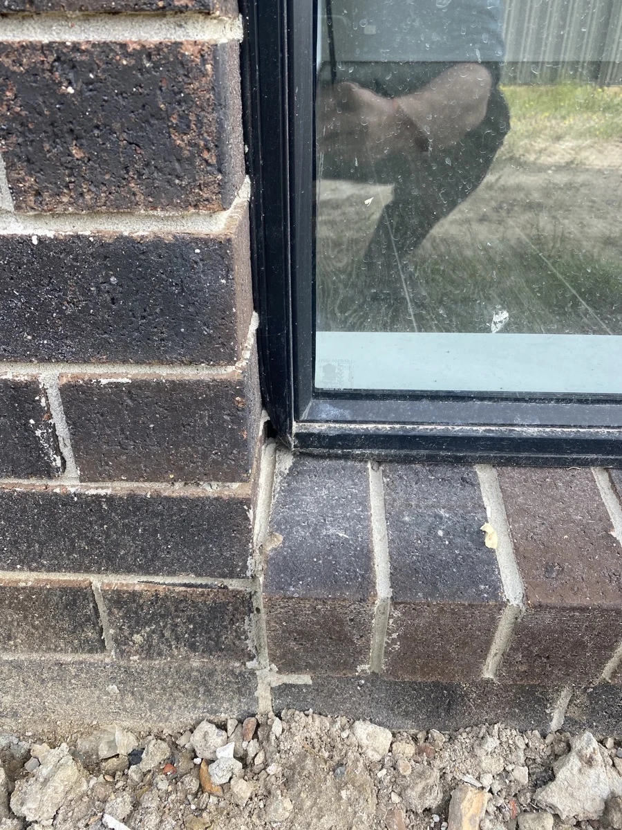 Close-up of a brick wall with a window frame. Dark bricks and dirt visible below. Reflection of a person in the window glass.