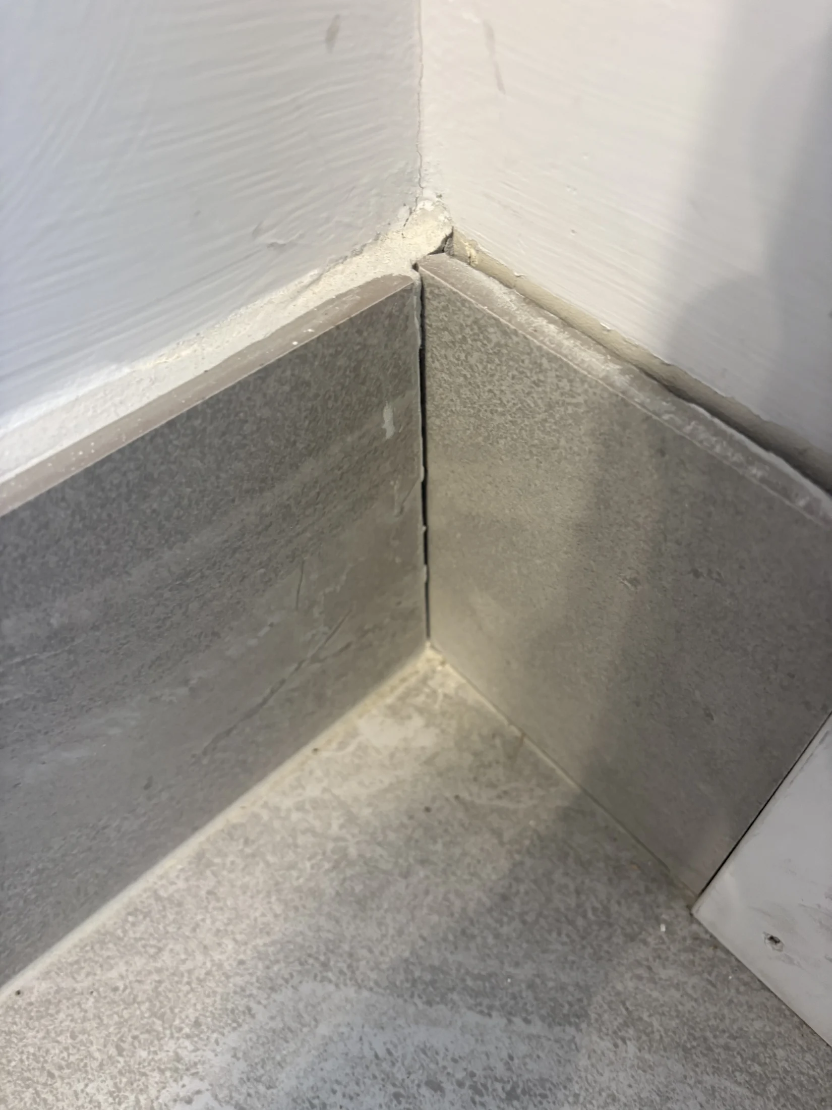 Corner of a room with two grey tiles meeting on the floor and a white painted wall above. The corner is filled with caulk.