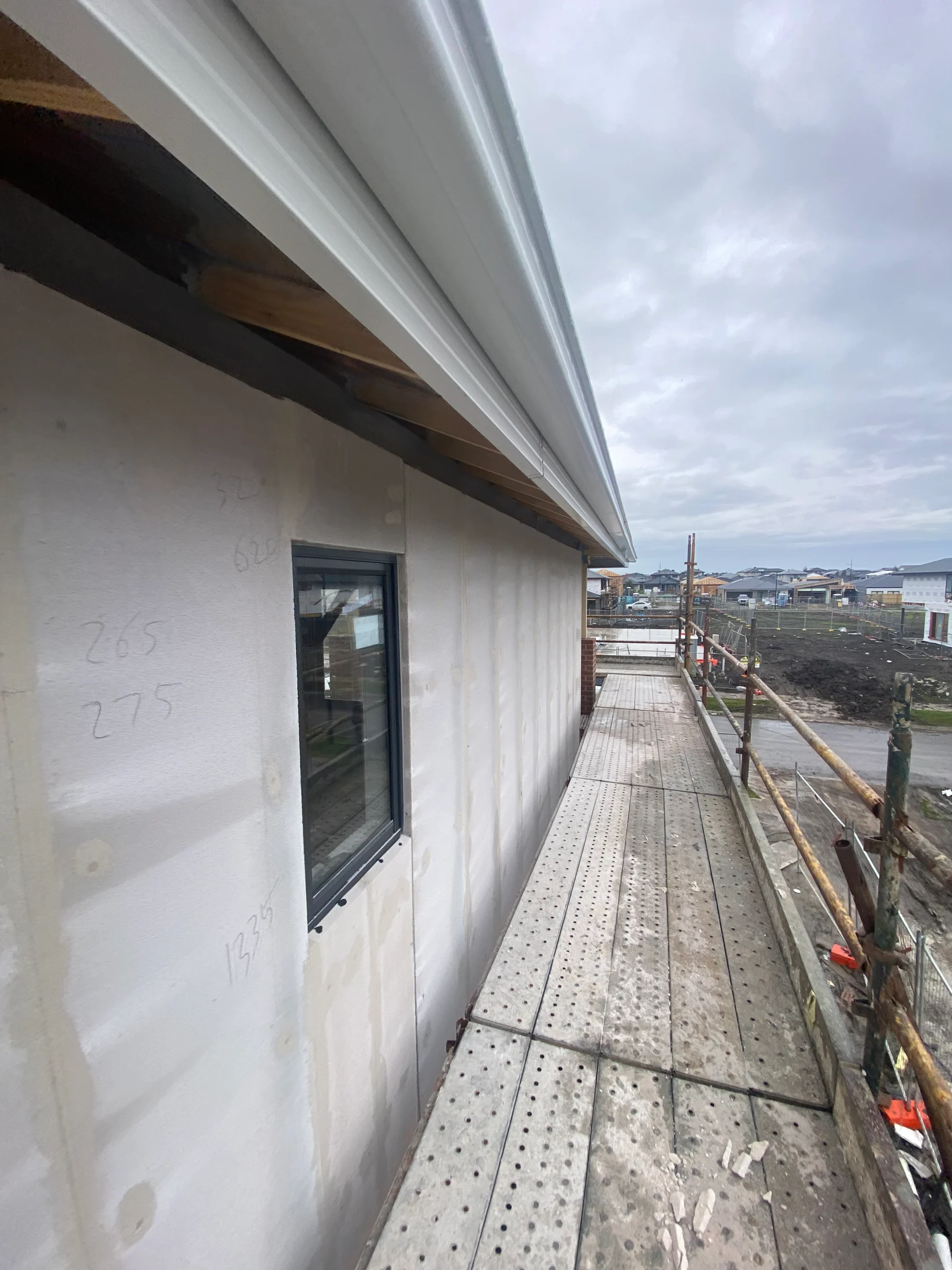Construction site with scaffolding beside a white building. Overcast sky, visible handwriting on the wall. Urban landscape in the background.