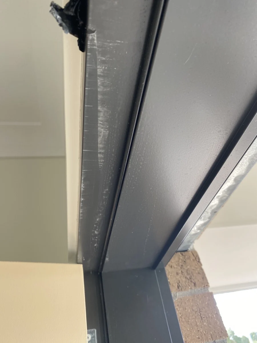 Close-up of a black metal door frame with visible scratches. Beige wall and brickwork form the background. Bright lighting highlights textures.