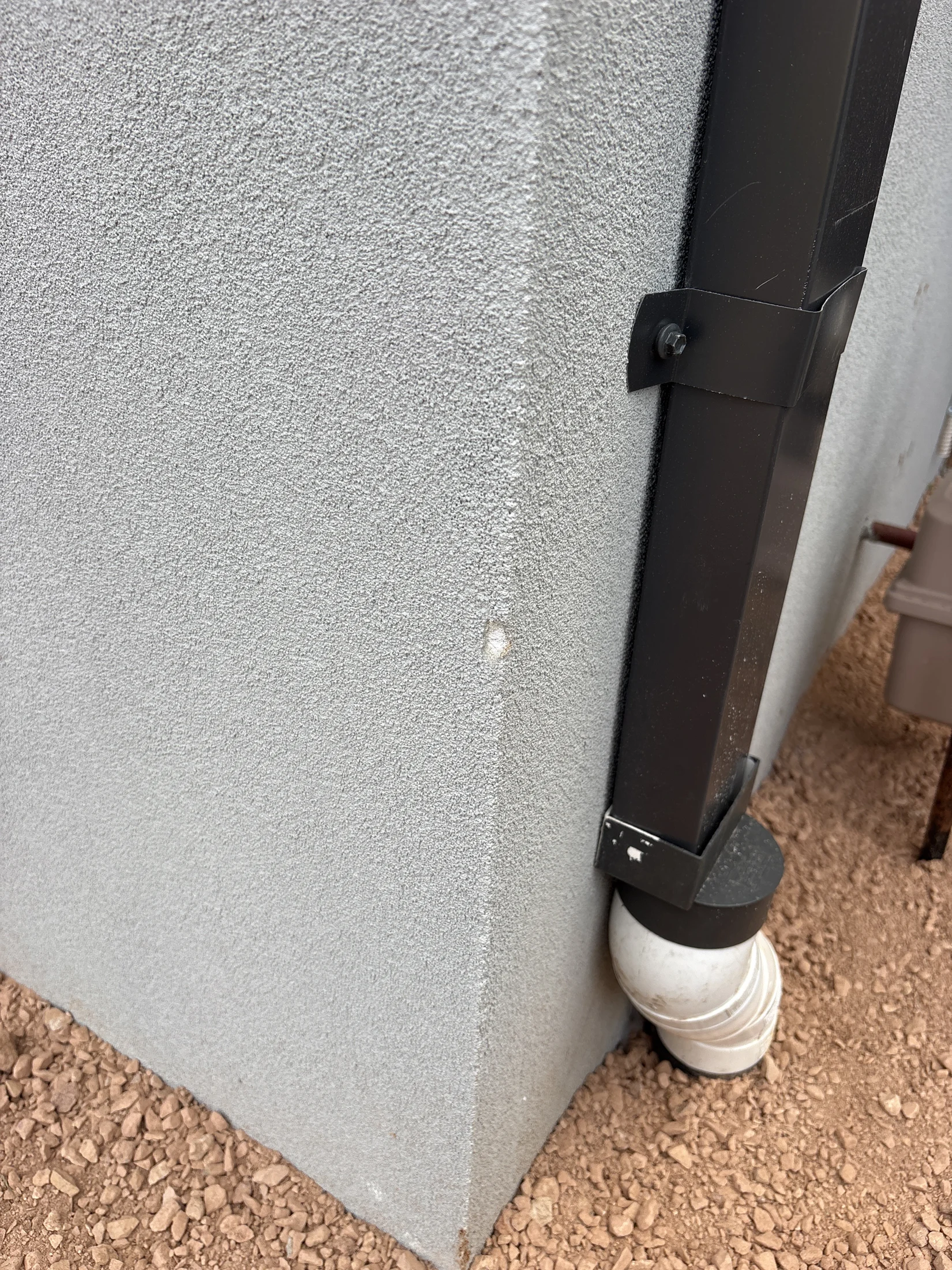 Corner of a stucco wall with a black metal drainpipe. The wall is light gray with a small chip. The ground is covered in brown gravel.