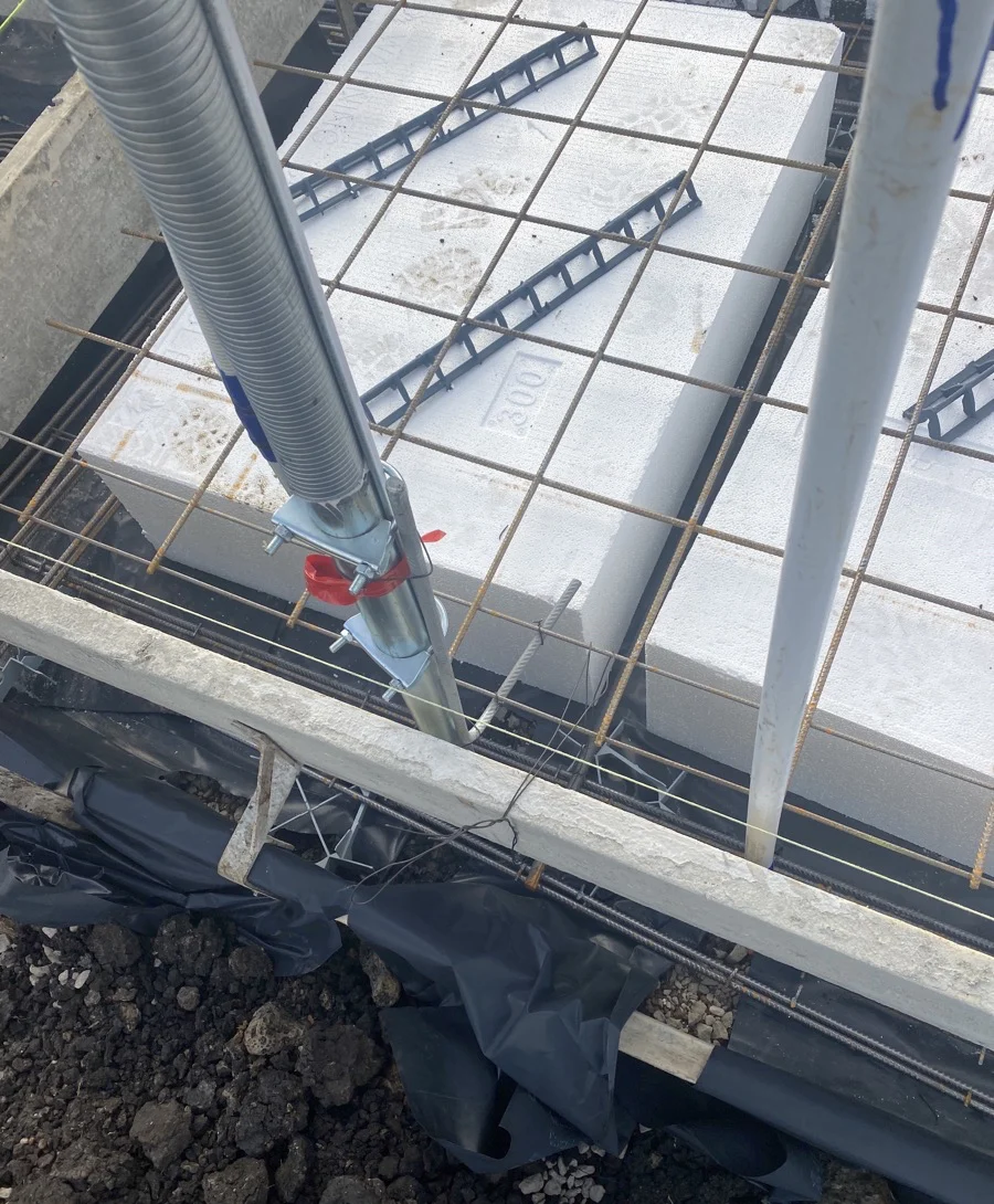 Construction site view showing steel bars over white blocks, labeled "100," with black plastic sheets and gravel beneath.