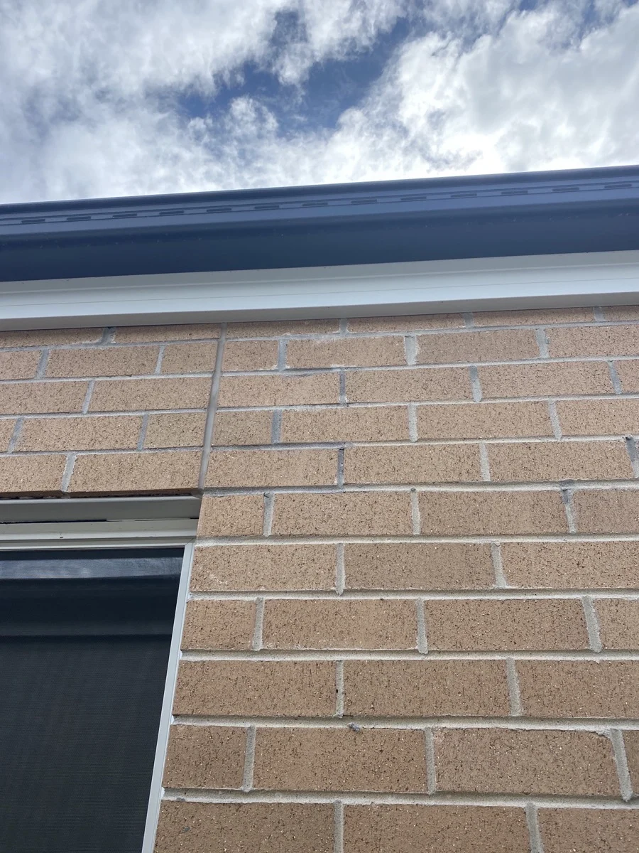 Brick wall with a dark window in the lower left, beneath a gutter. Blue sky and clouds are visible above, creating a calm scene.