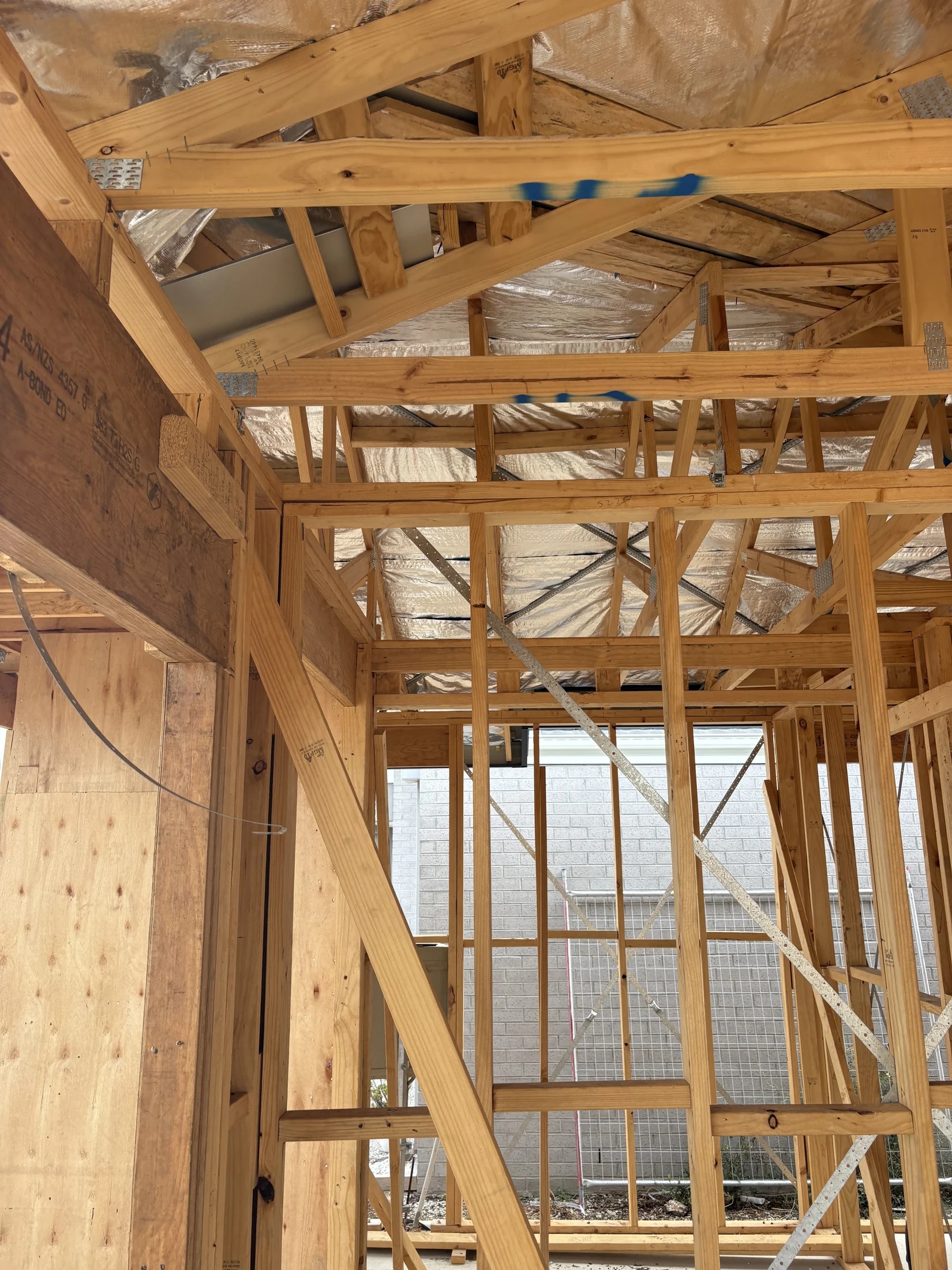 Wooden framework of a house under construction, with exposed beams and metal braces. Background shows a brick wall. Natural light illuminates the scene.