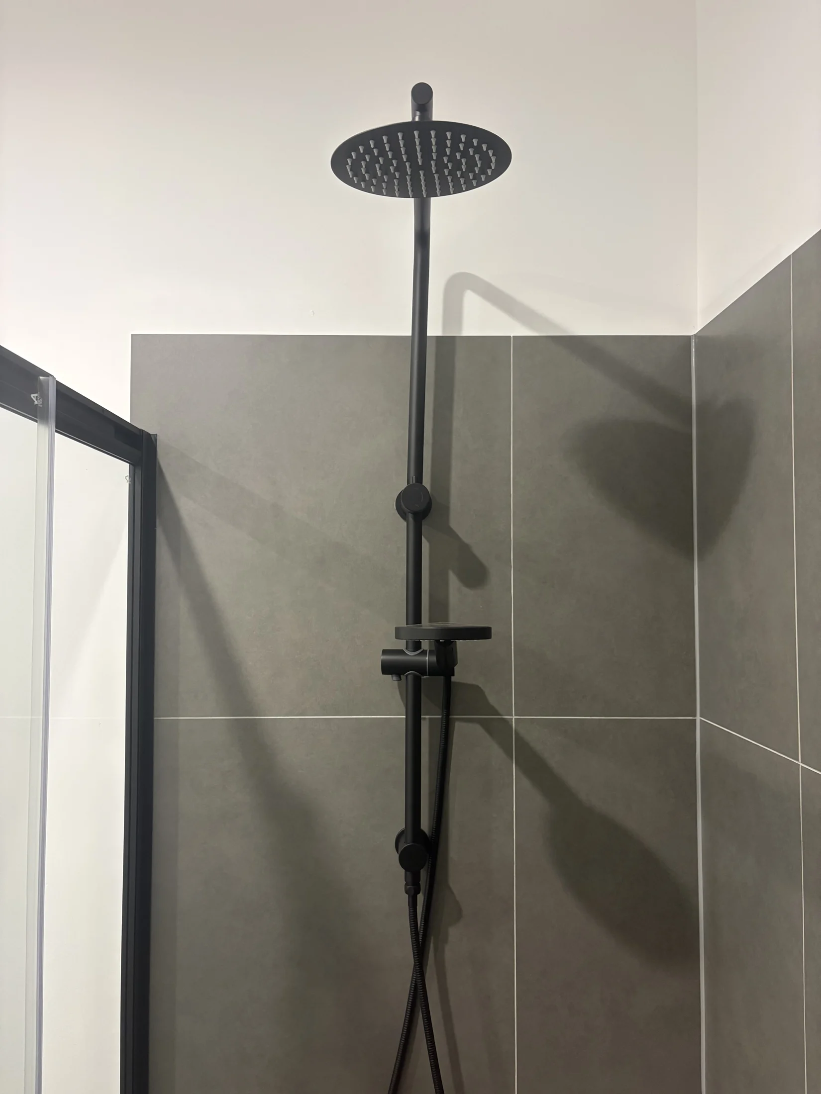 Black rain showerhead and control panel against gray tiles in a modern bathroom. Glass shower door partially visible. Minimalist design.
