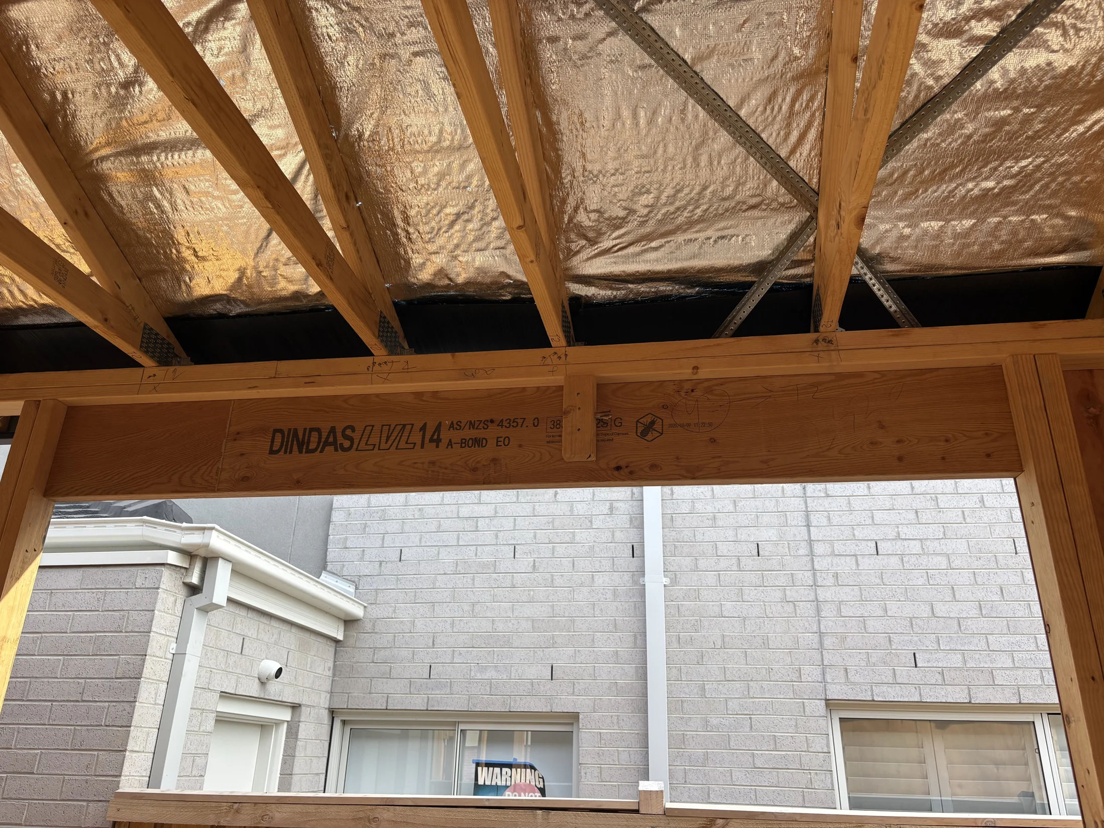 Wooden beams with "DINDAS LVL 14" text under a foil-lined roof. White brick wall and window visible in the background. Construction site.