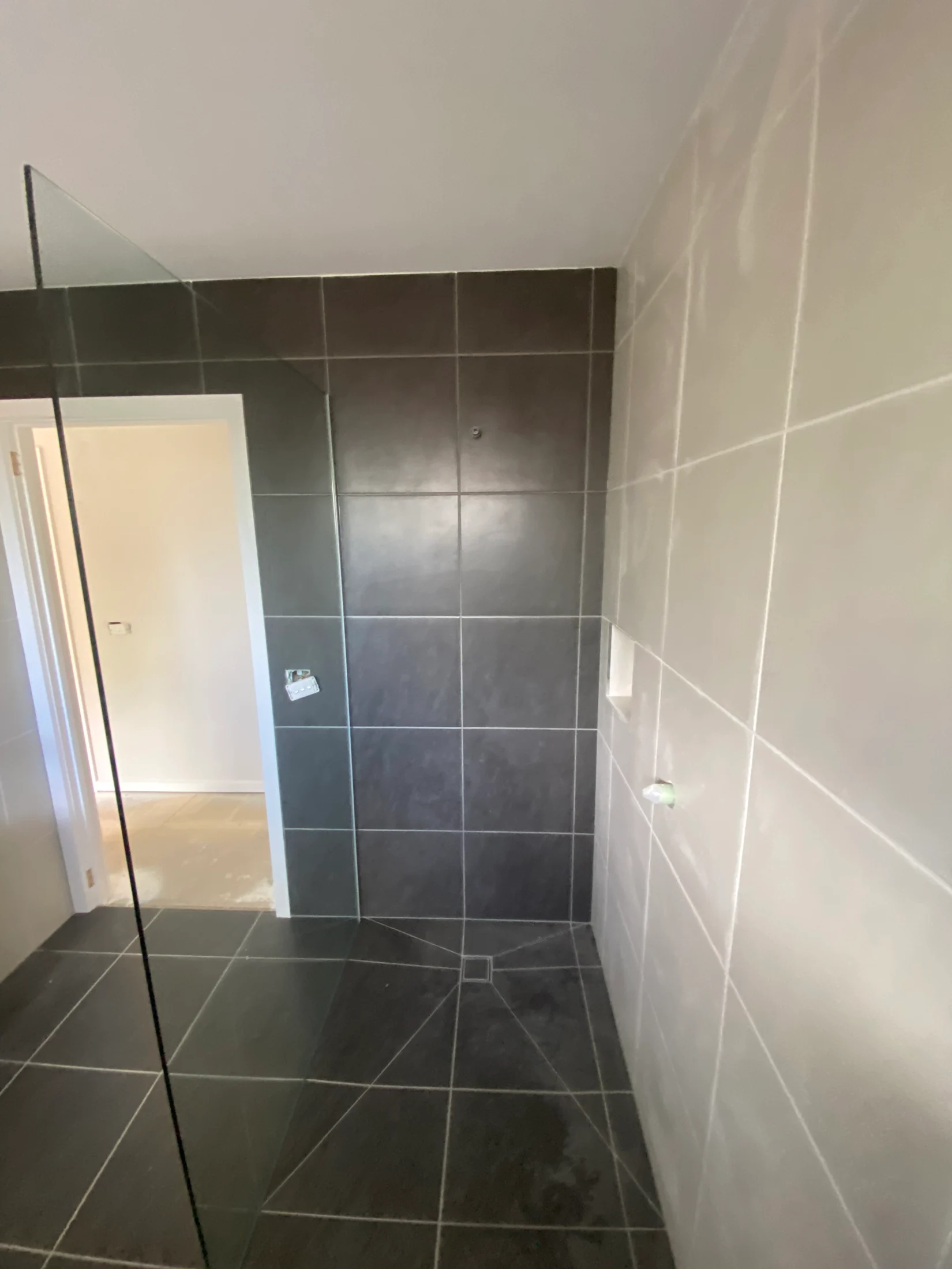 Shower area with dark gray tiles, glass partition, light gray walls, and towel hook. Doorway visible leads to a brighter room.