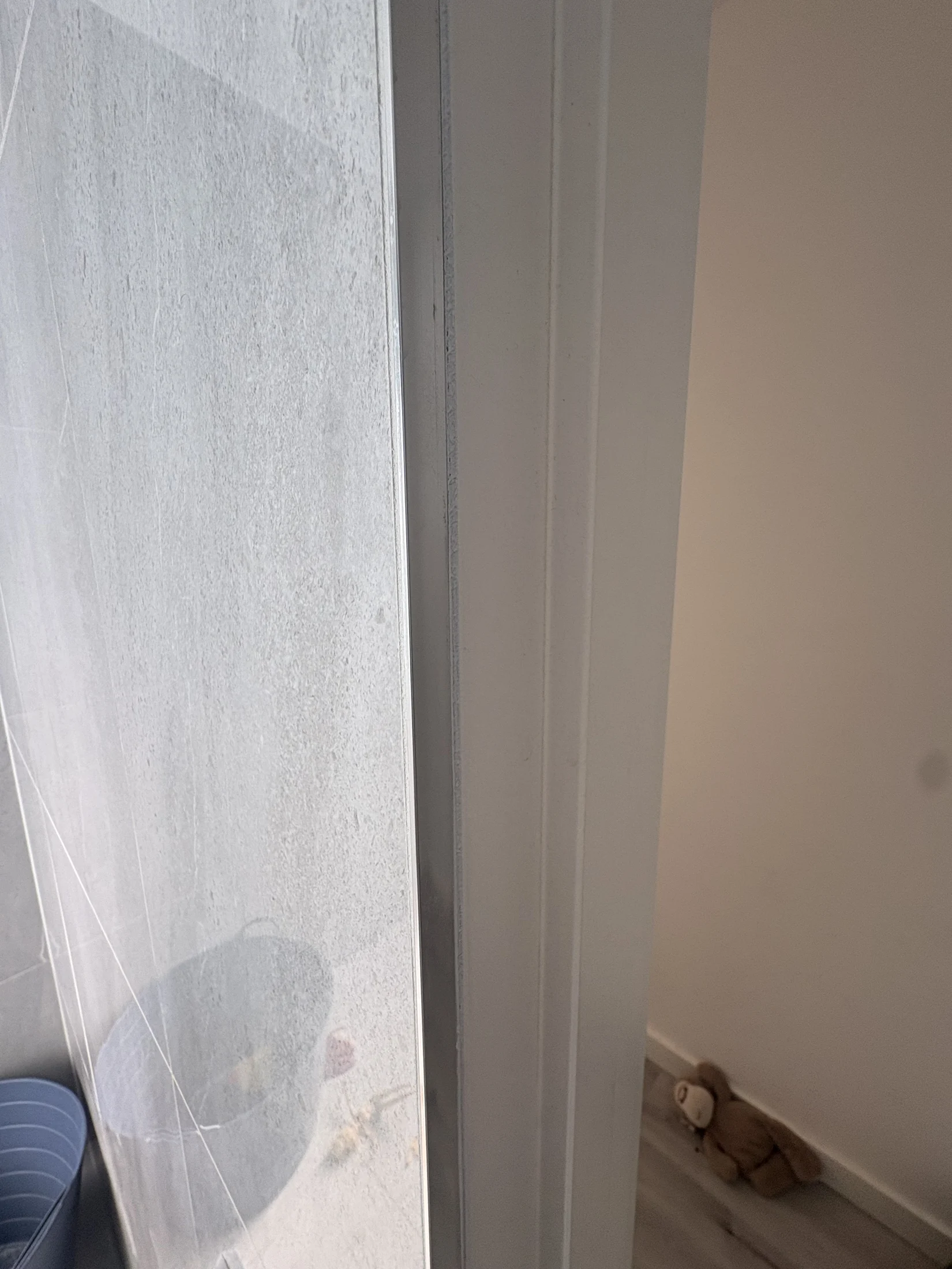 A teddy bear lies on a light wooden floor near a white wall. A frosted glass door partially reveals a blue and gray container.