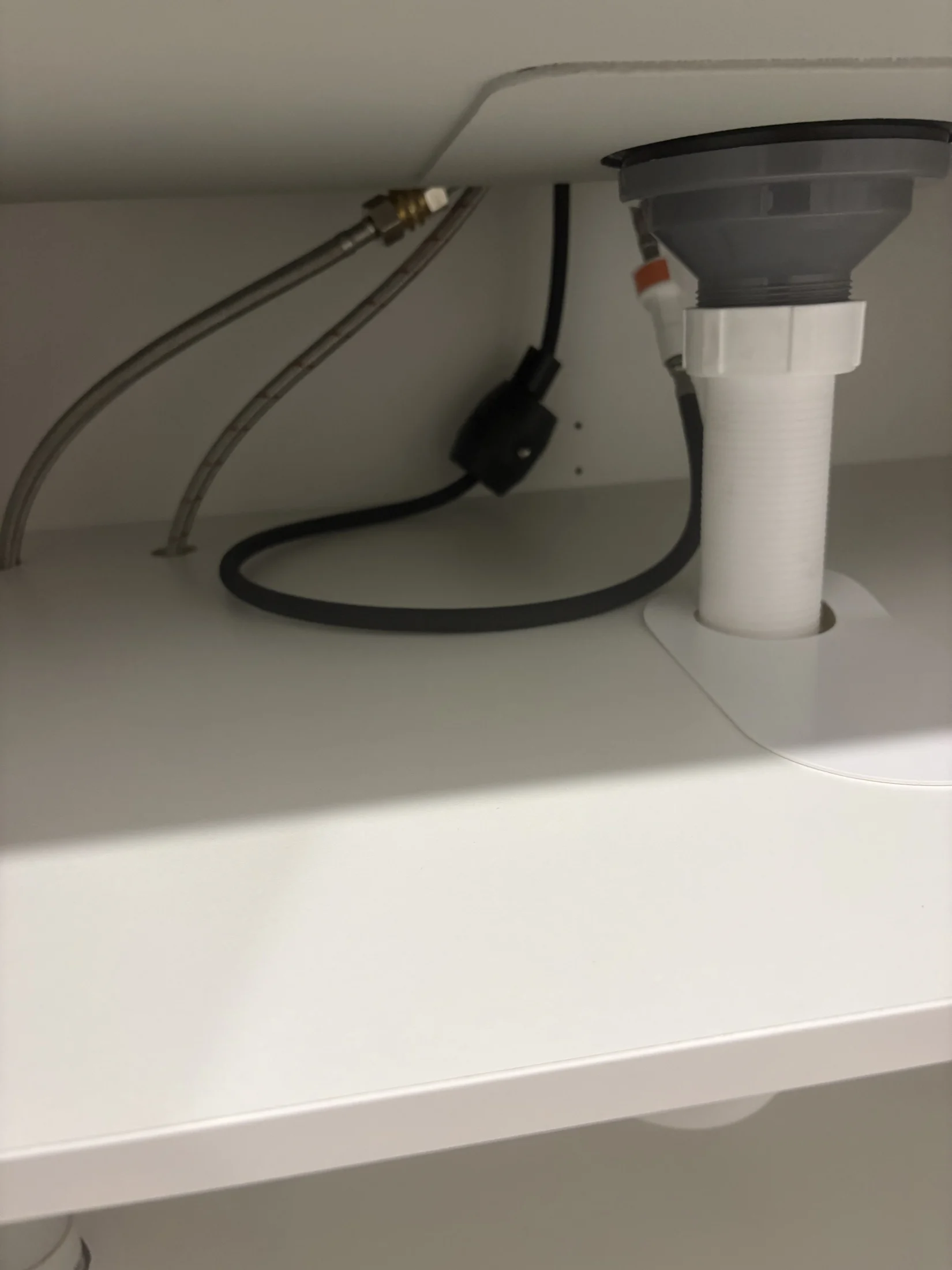 Under-sink view showing plumbing pipes and cables against a white shelf. No text or notable action visible. Neutral mood.
