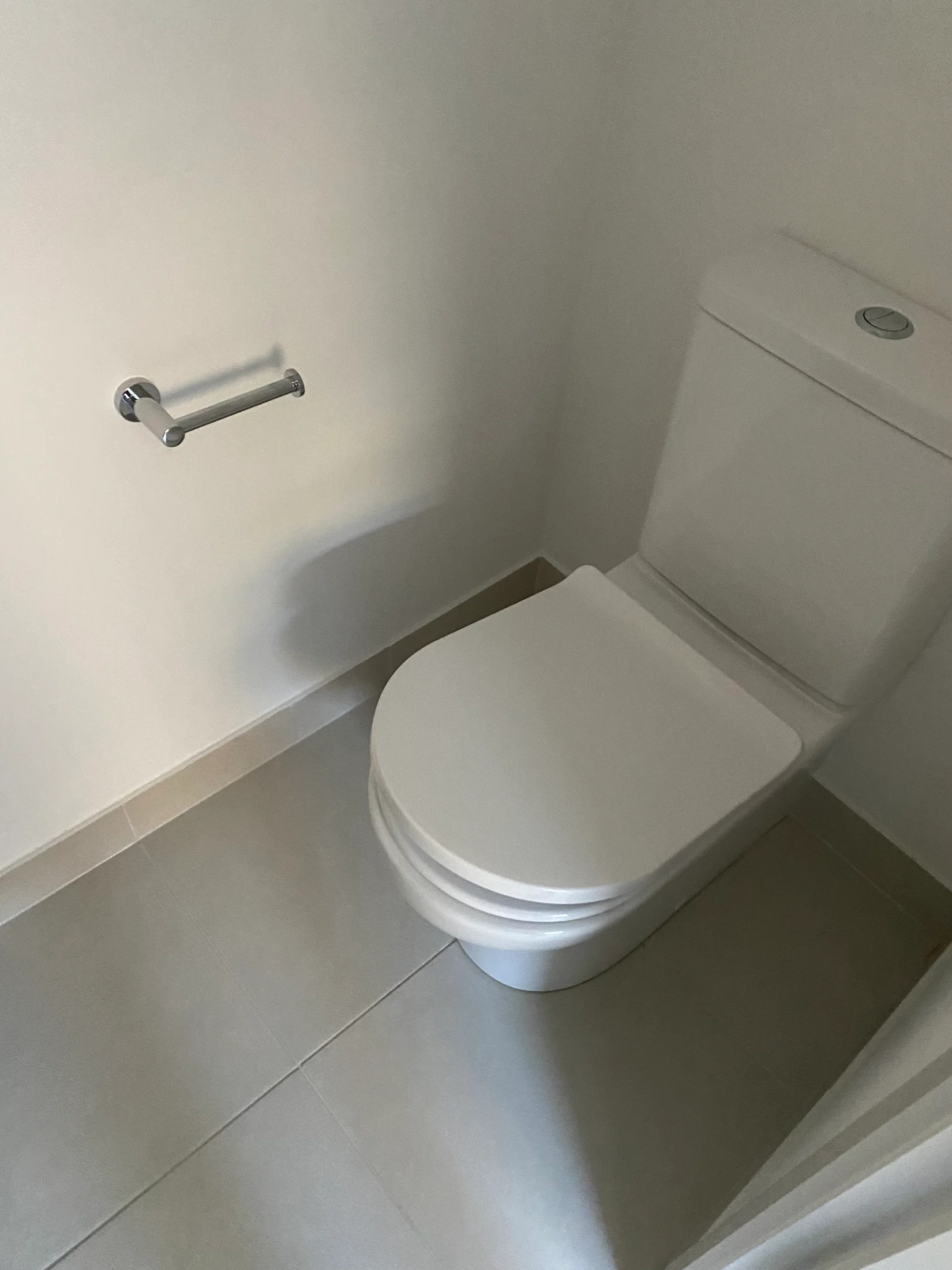 White toilet with closed lid in a small bathroom. Light gray tile floor, empty chrome toilet paper holder on the wall.