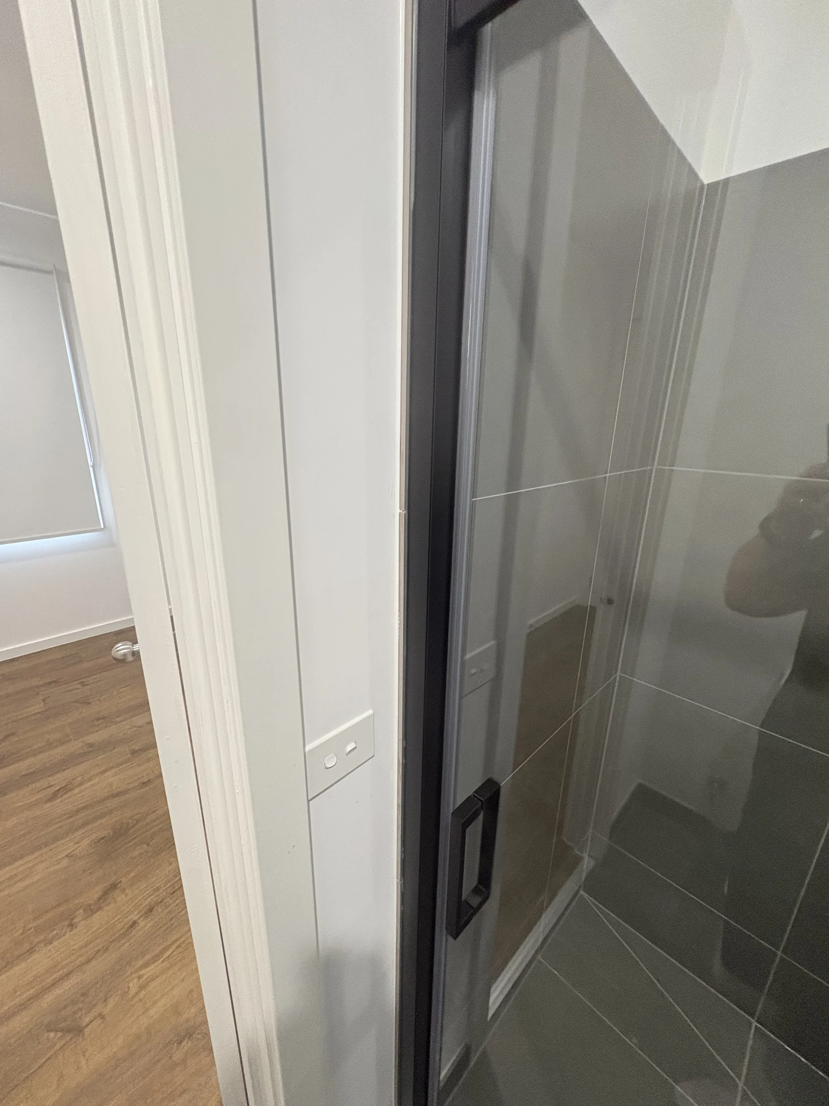 Bathroom corner with glass shower door and black frame. Light switches on white wall, wooden floor, and window with blinds in background.