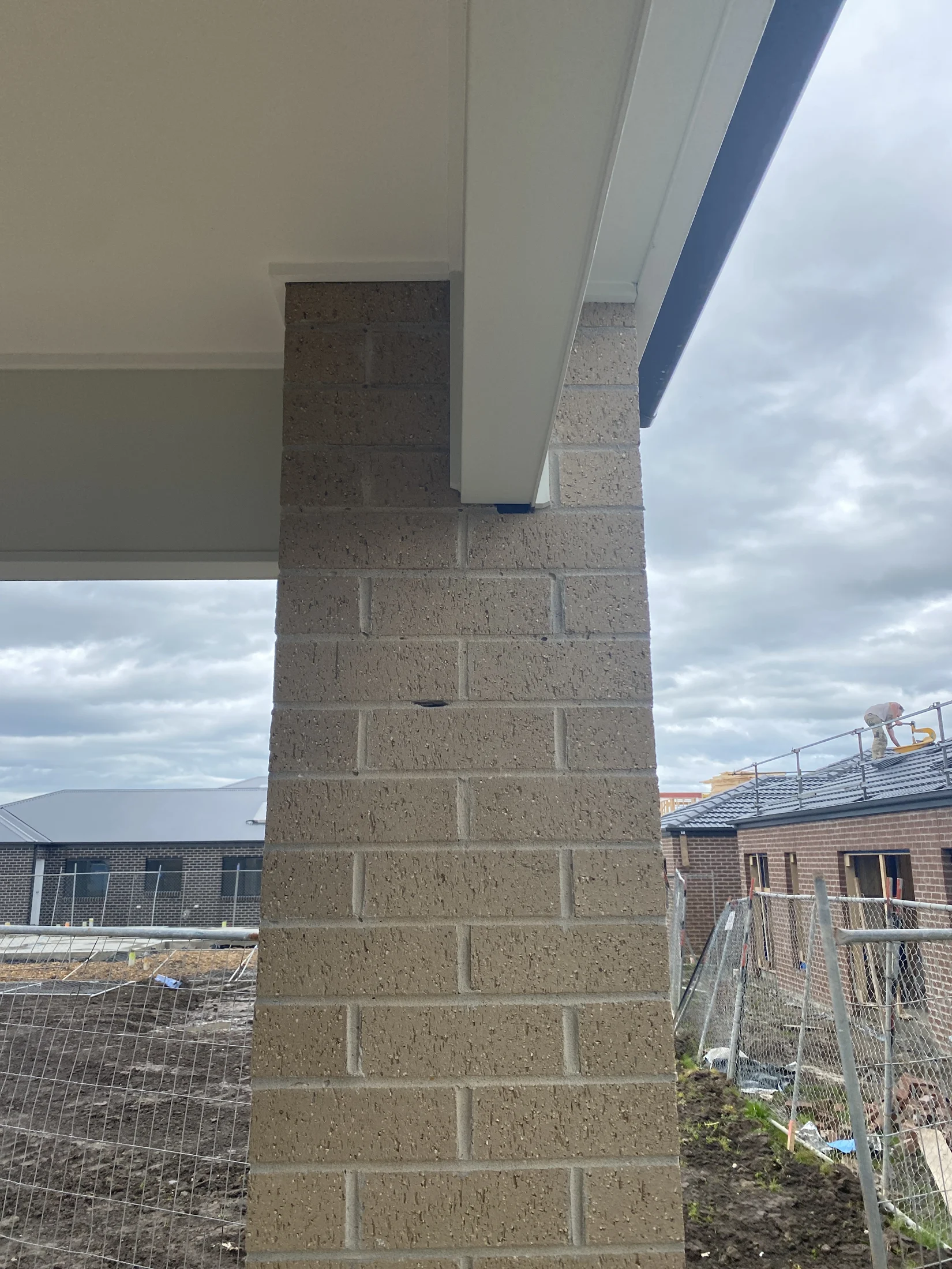 Brick pillar supports a roof against a cloudy sky. In the background, houses under construction with scaffolding and dirt ground.