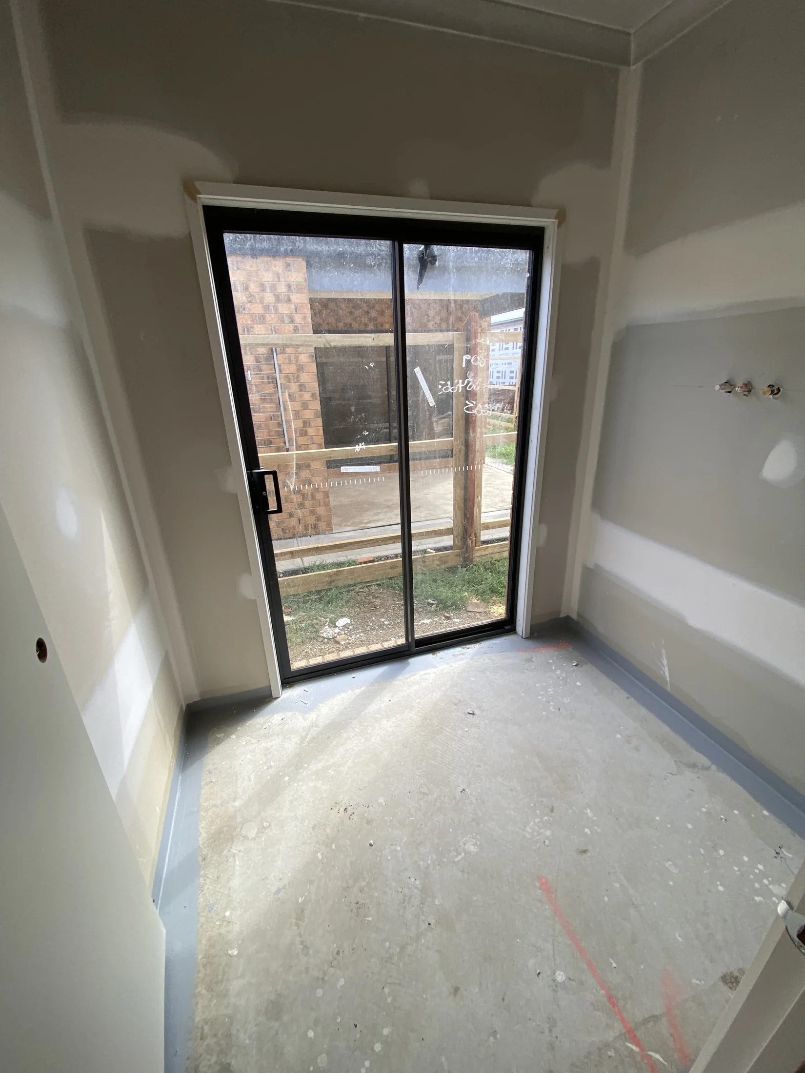 Unfinished room with drywall, concrete floor, and a glass sliding door showing a brick wall outside. White scribbles on the glass.
