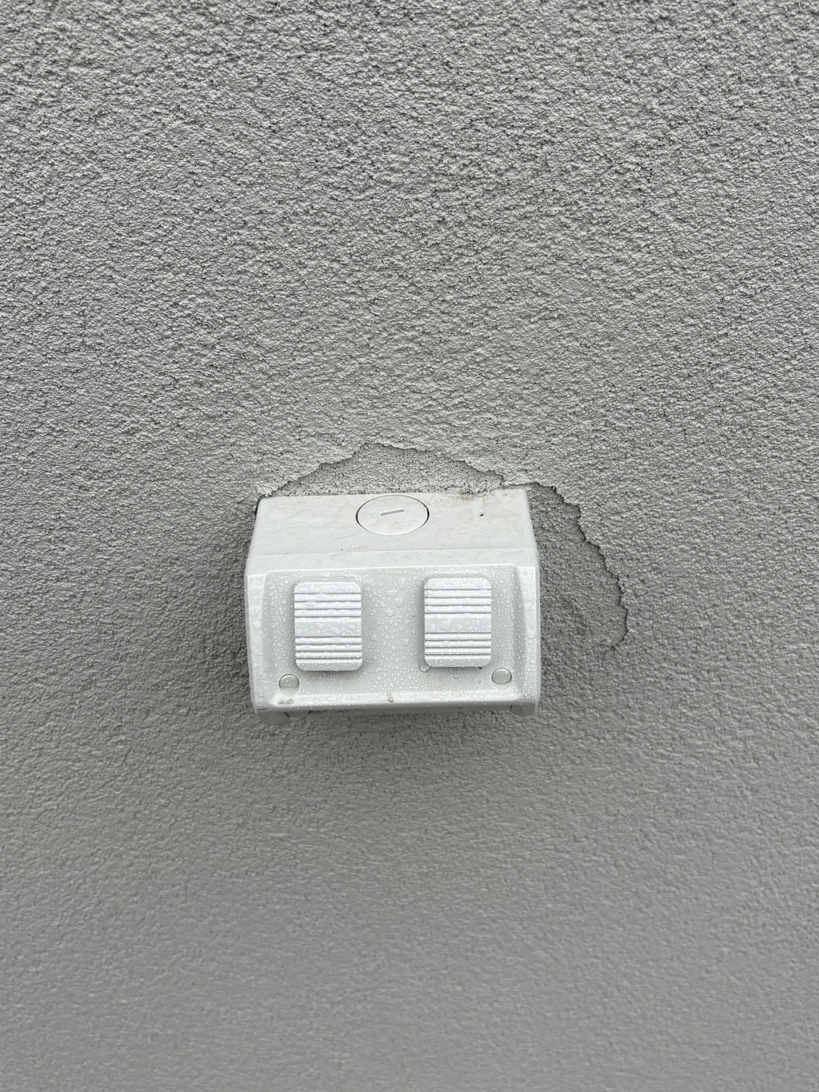 Gray textured wall with a white dual outdoor electrical outlet cover, damp with rain. Peeling paint visible around the cover.