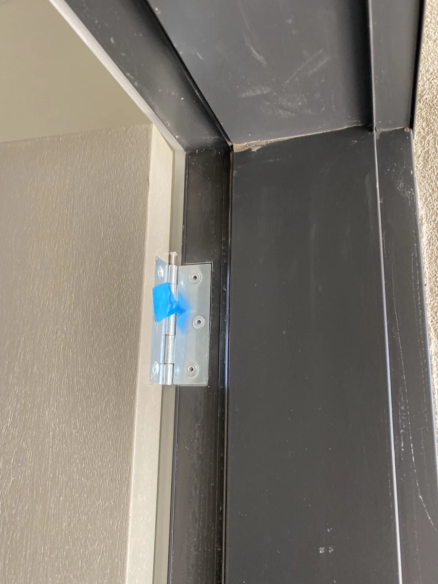 Door hinge secured with blue tape on a dark frame. Beige wall in the background. No visible text, minimalistic and neutral setting.
