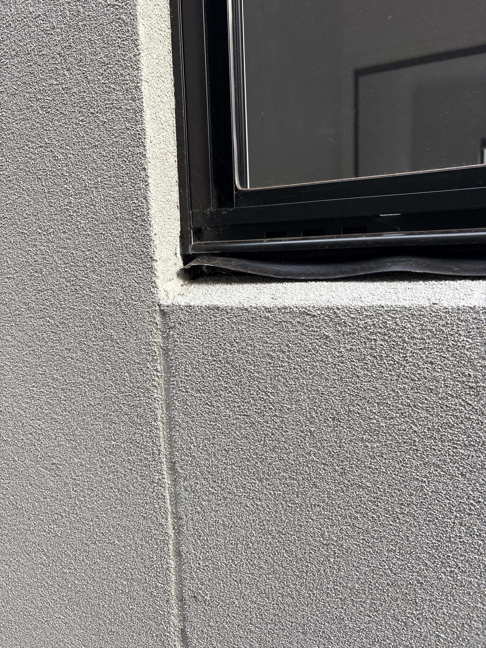 Close-up of a black-framed window on a textured gray wall, showing a gap at the bottom edge. No visible text or significant actions.