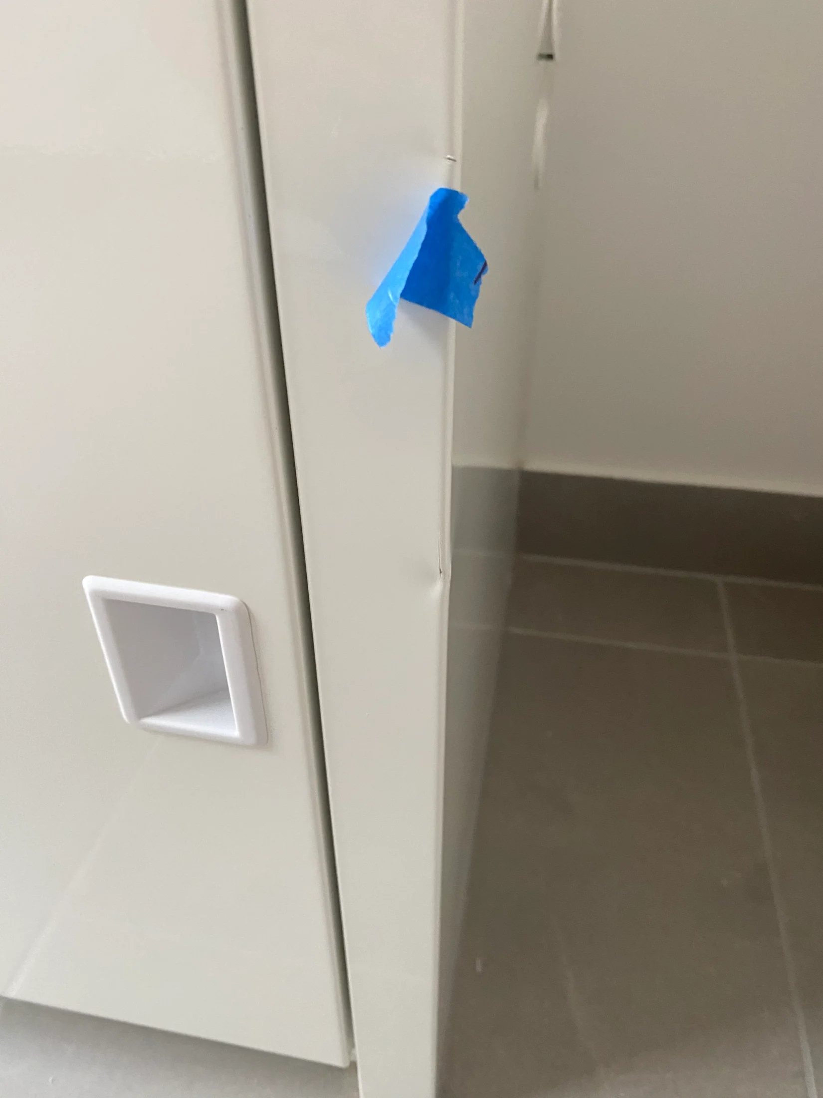 White cabinet in corner of room with a small dent marked by blue tape. White square handle and gray tiled floor visible.