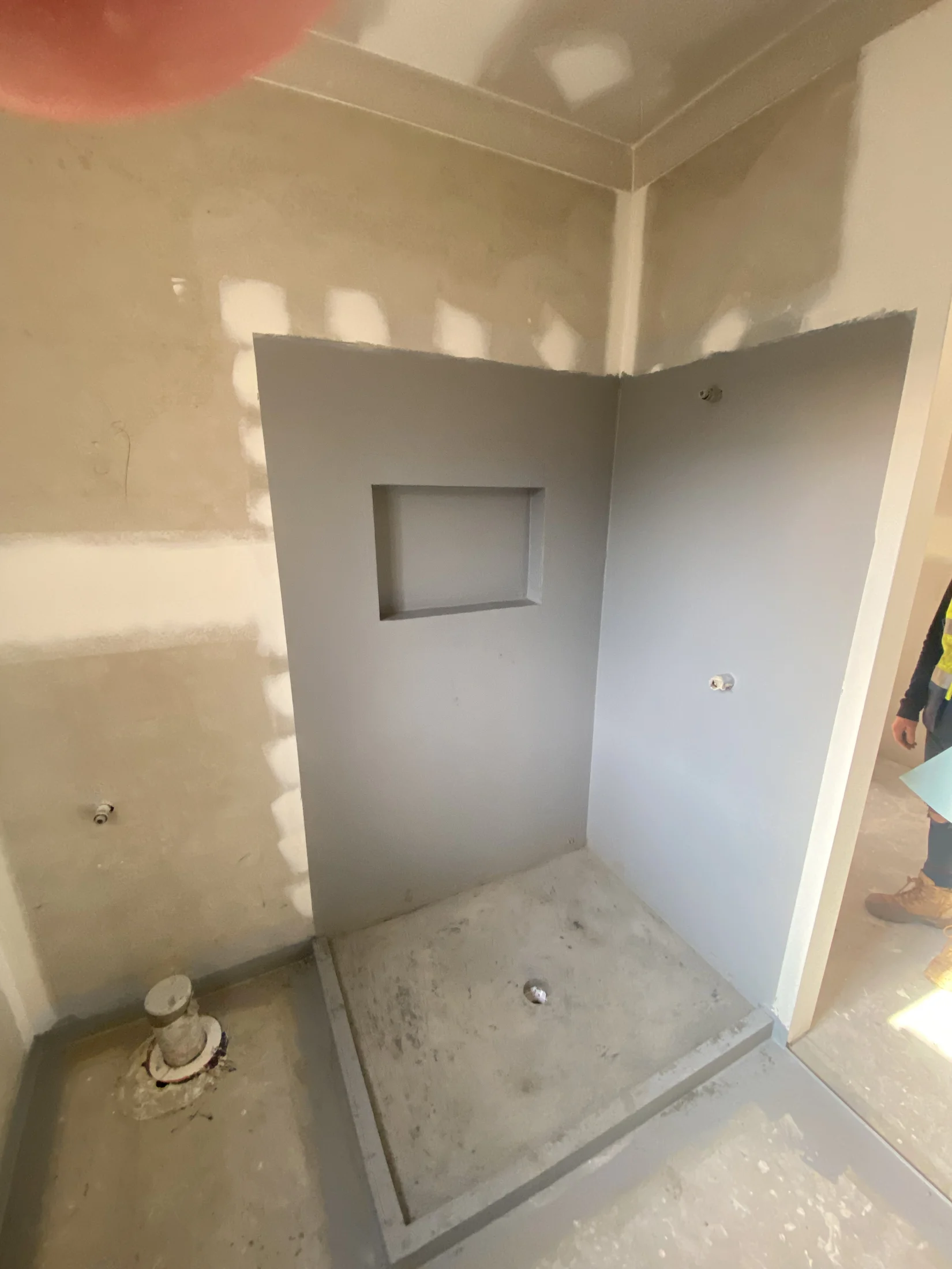 Unfinished bathroom with a grey shower alcove, drain, and partial plumbing. A person in work boots is visible, suggesting ongoing construction.