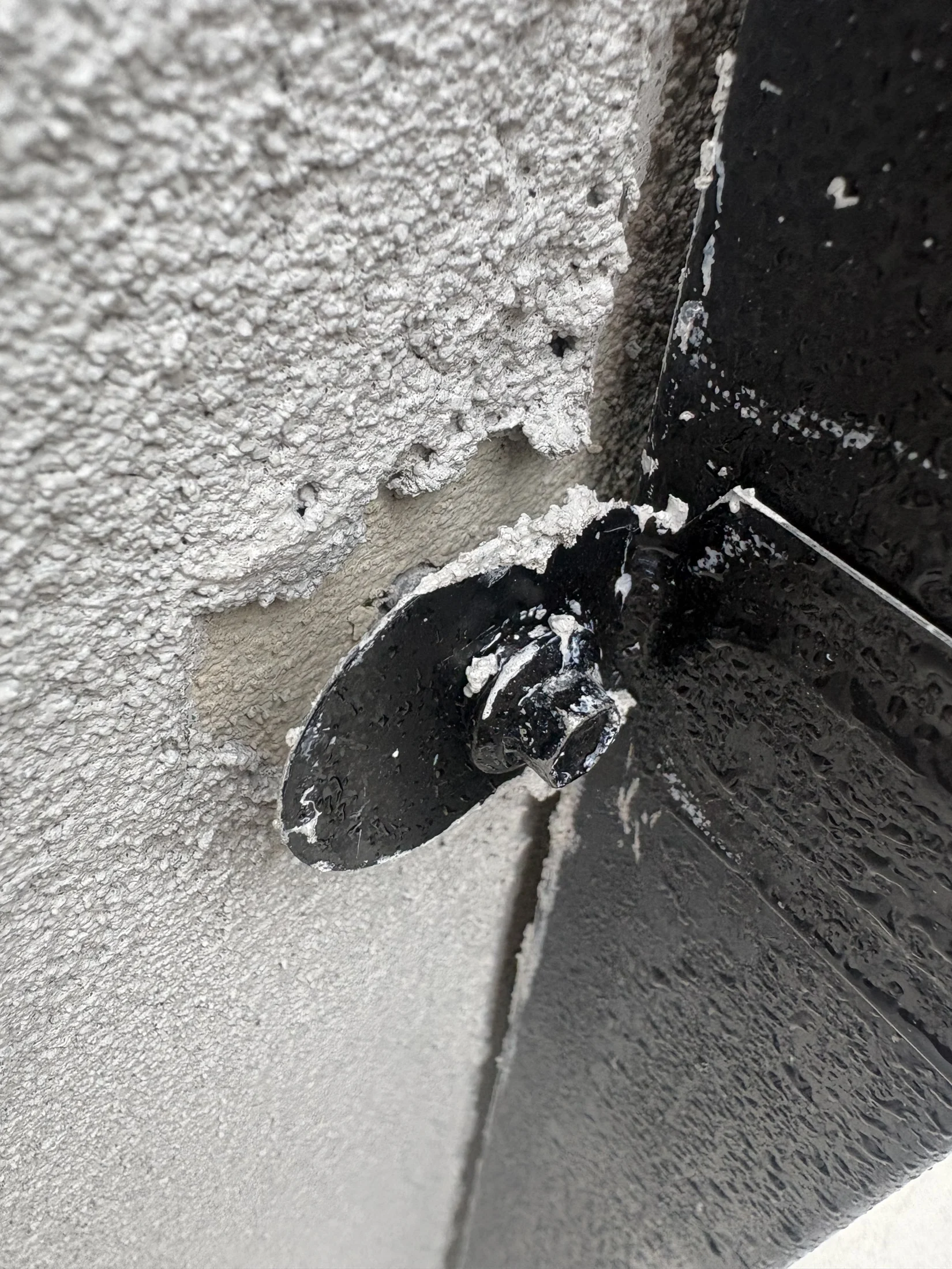 A close-up of a black metal bolt on a textured white wall with some chipped areas, revealing underlying material. The scene is detailed and weathered.