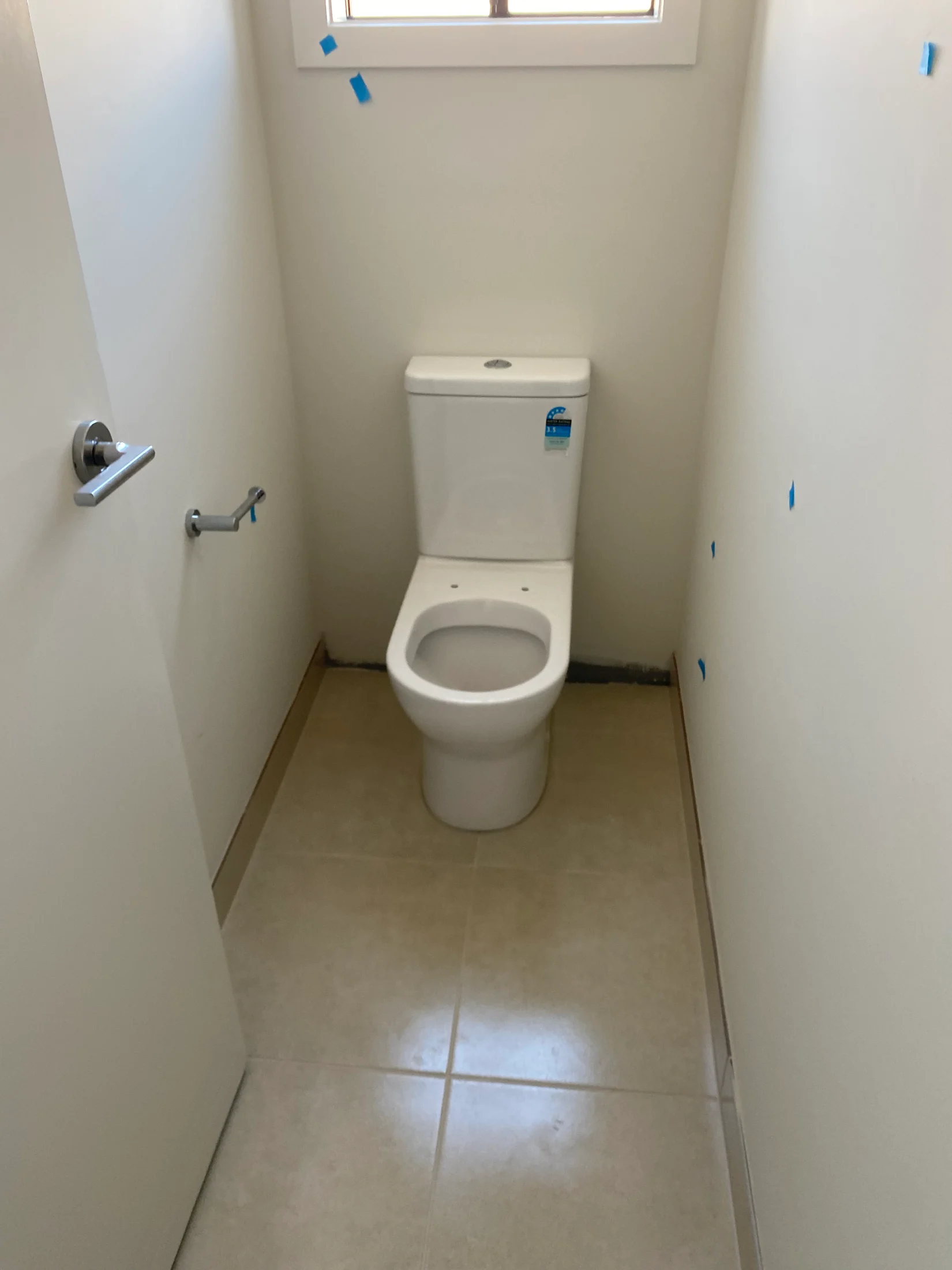 A white toilet in a small, tiled bathroom. Blue tape is on the walls for marking. The door is ajar with a silver handle.
