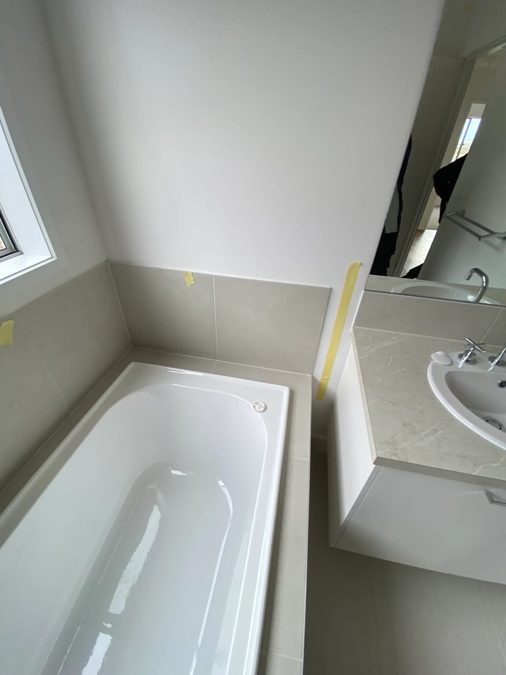 Bathroom with a white bathtub, beige tiles, white sink on a countertop, and a small window. Yellow tape is visible on the walls.